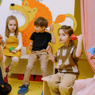 A teacher and young children play with colorful bells in a cheerful classroom with animal-themed decor—an example of how to teach your children well through creative play and engagement.